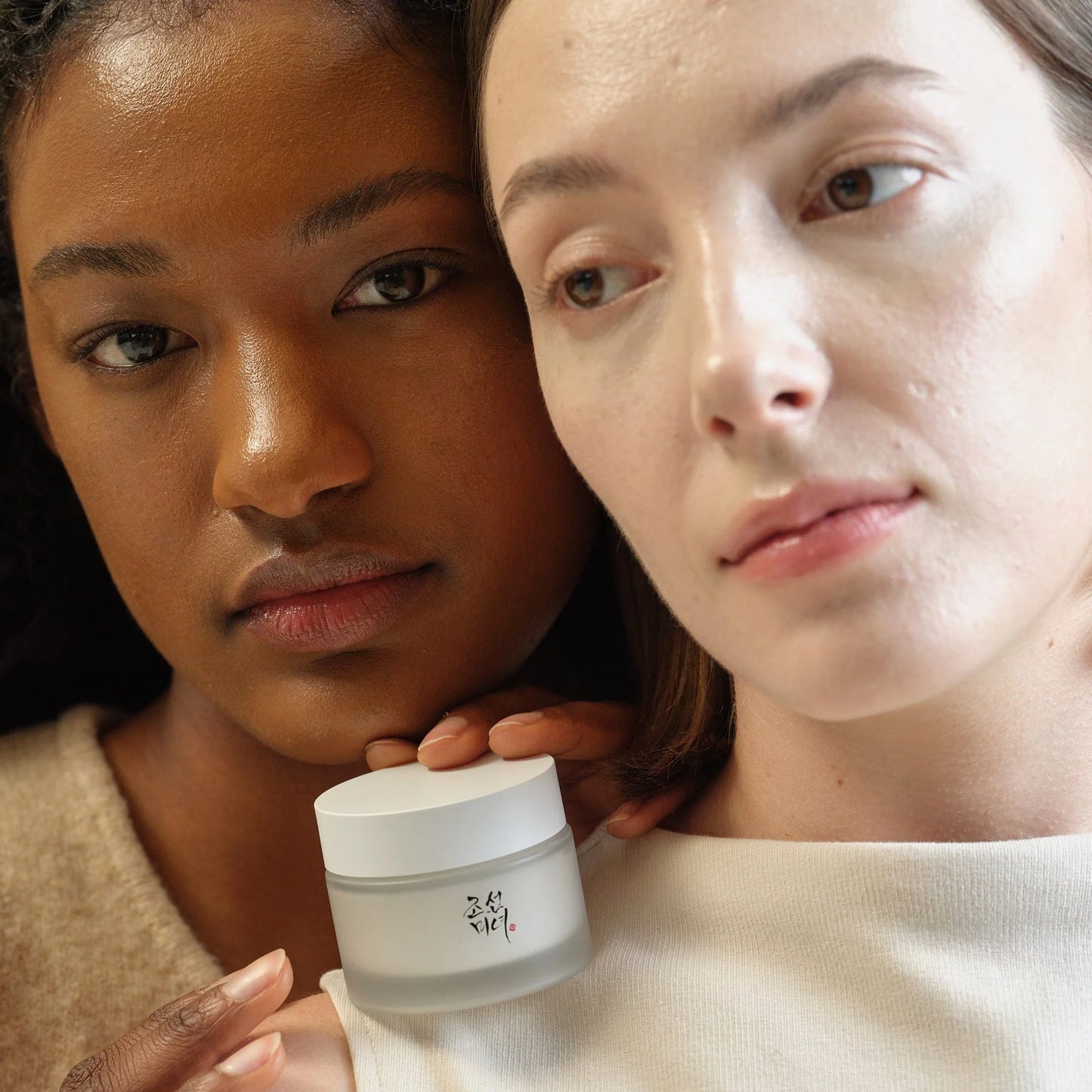 Zwei Frauen, eine hält eine helle Cremedose. Beide haben neutrale Gesichtsausdrücke.