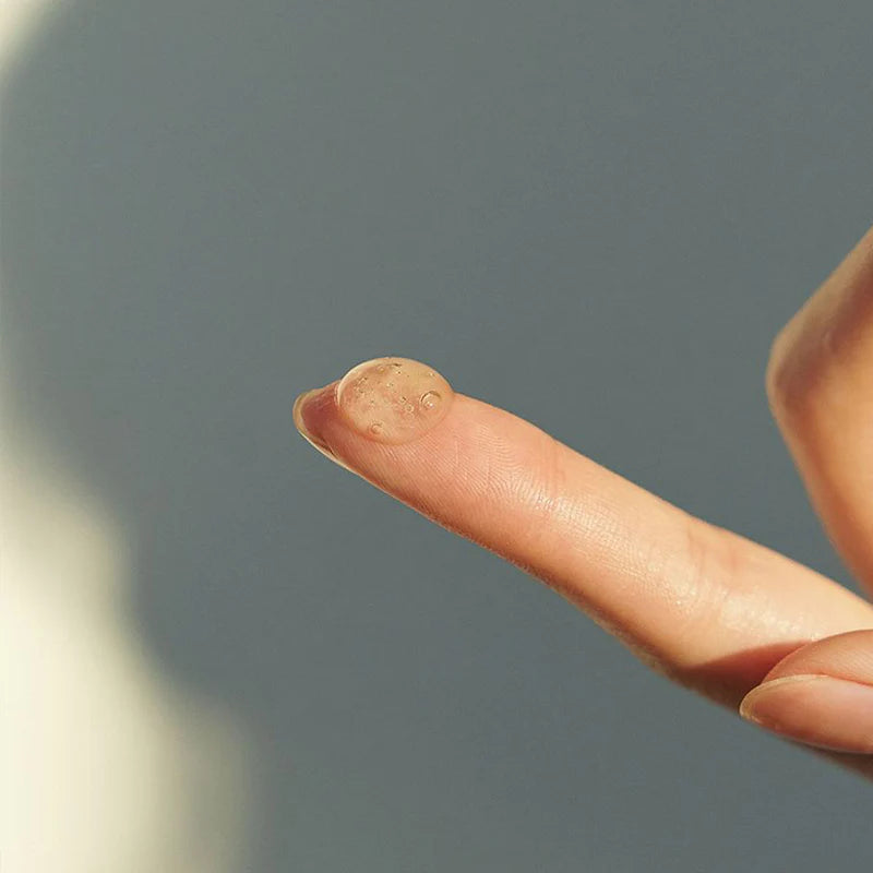 Ein Finger zeigt einen Tropfen des Serums, grauer Hintergrund.