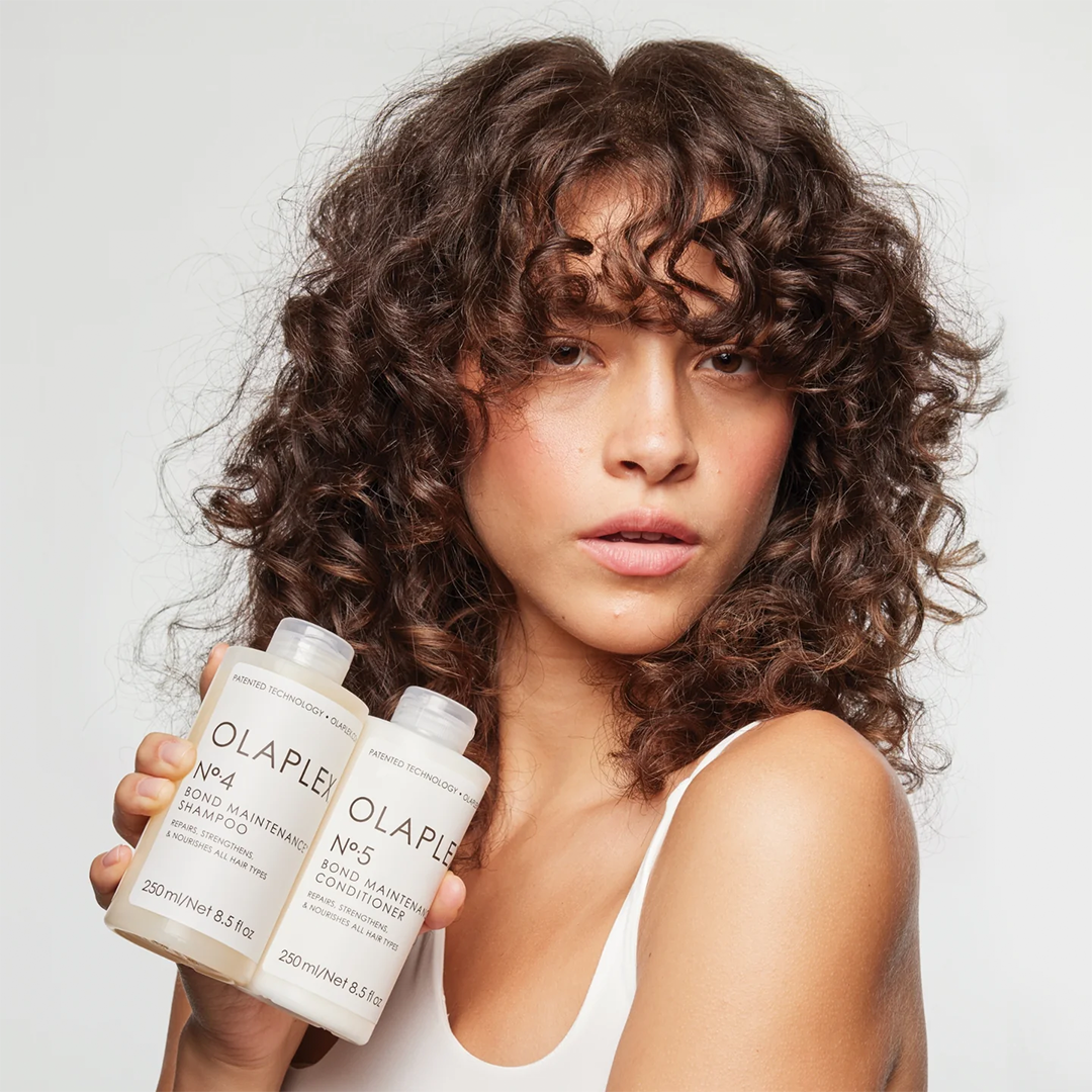 Frau mit lockigem Haar hält Olaplex No. 4 Shampoo und No. 5 Conditioner.
