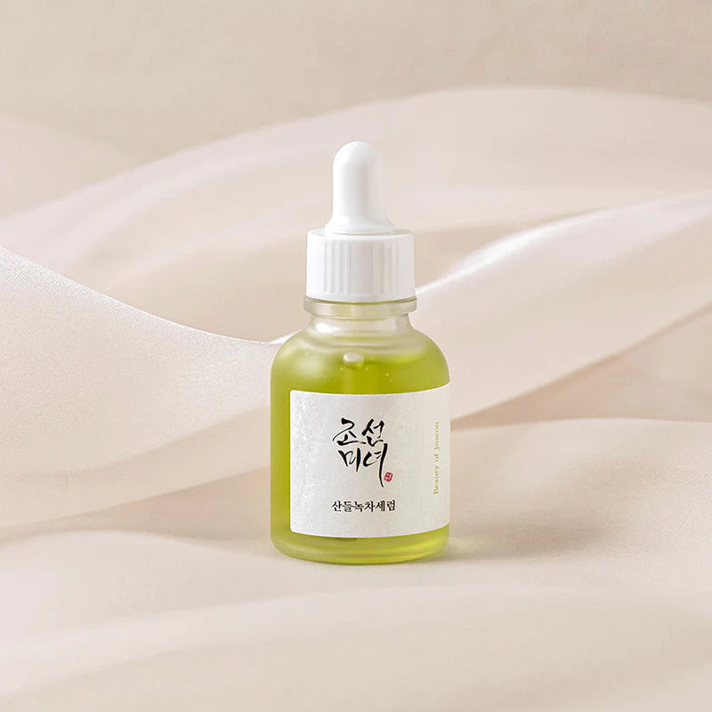 Eine kleine Flasche mit grünlichem Serum und weißer Pipette, auf einem drapierten beigen Stoff.
