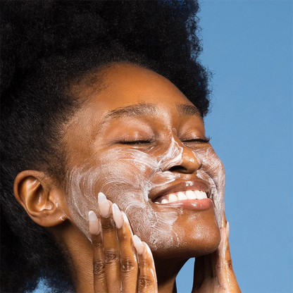 Frau mit aufgeschäumtem Cleanser im Gesicht, lächelnd vor blauem Hintergrund, zeigt Freude und Pflege.